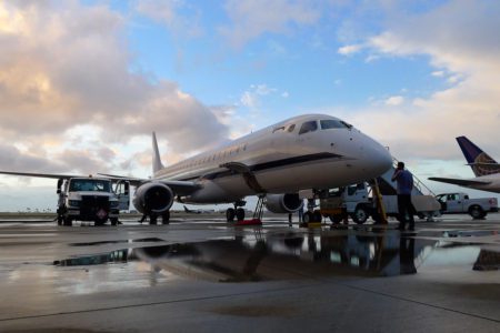 A private jet parked on a tarmac, surrounded by ground crew and service vehicles, with a cloudy sky reflecting on wet pavement.